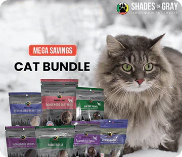 Shades of Gray Indigenous Pet Treats' Mega Cat Pet Treat Bundle, featuring a close-up of a majestic gray tabby cat with green eyes gazing curiously at the camera against a snowy outdoor background. In the foreground, a stack of colourful product packages showcasing the bundle's contents. The brand logo with a paw print is in the top left, and bold red 'MEGA SAVINGS' and 'CAT BUNDLE' text overlays the top right.
