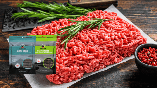 Raw ground bison with fresh rosemary, surrounded by bags of Shades of Gray Bison Rolls and Beaver Rolls novel protein dog treats.