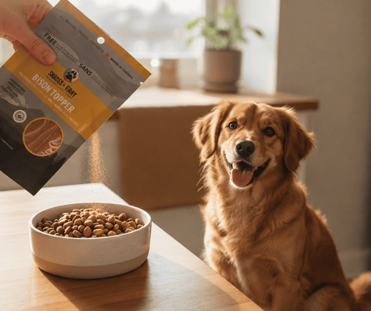 A hand holding a bag of Shades of Gray single-ingredient meat topper over a bowl of kibble, while a happy golden retriever sits at the table waiting for dinner.