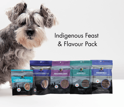 Dog looking at a lineup of Shades of Gray Indigenous Pet Treats on a white background, featuring Indigenous Strips, Bison Rolls, Venison Rolls, Rabbit Ears, and Freeze-Dried Bison Liver from the Indigenous Feast & Flavour Pack.