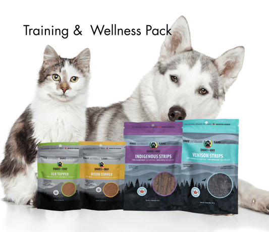 Dog and cat sitting behind a lineup of Shades of Gray Indigenous Pet Treats on a white background, featuring Indigenous Strips, Venison Strips, Bison Topper, and Elk Topper from the Health & Wellness Pack.