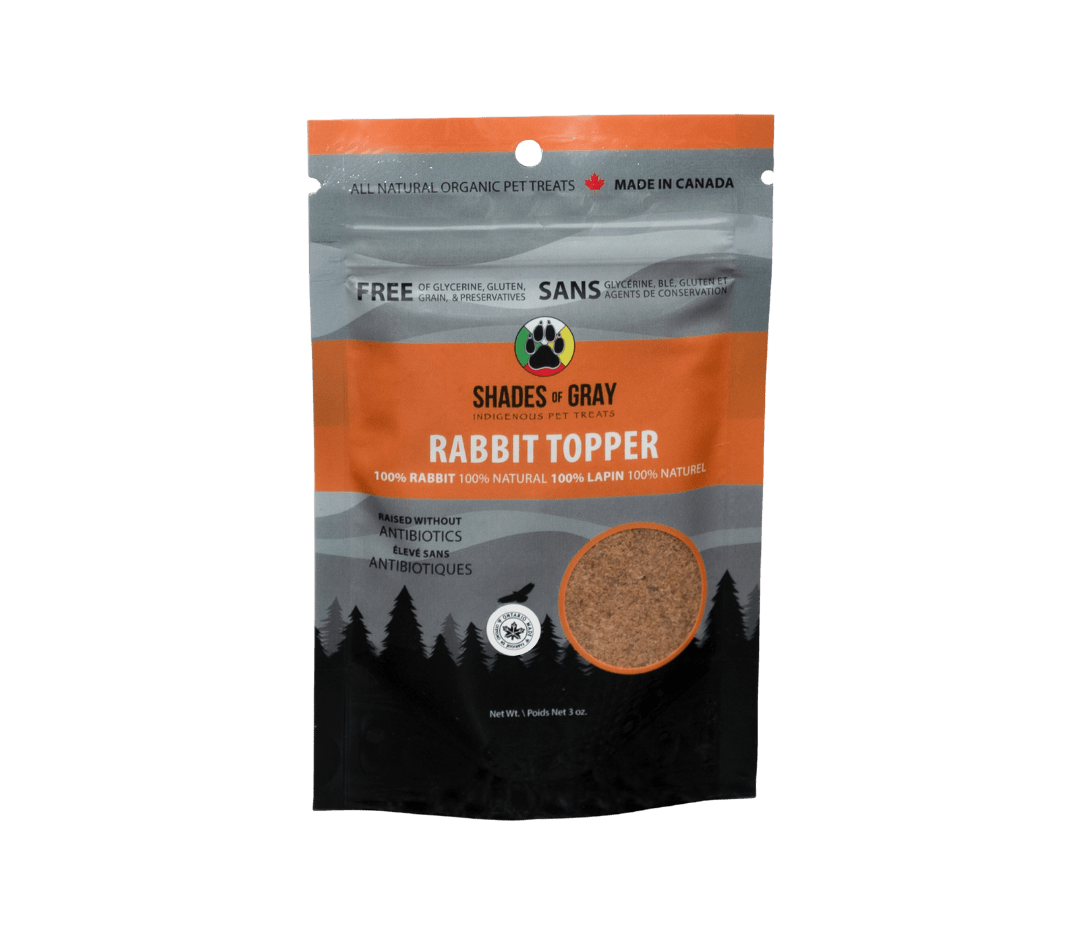 Front of Shades of Gray Indigenous Pet Treats Rabbit Topper packaging in a sleek orange and grey resealable pouch, featuring bold white text "RABBIT TOPPER," single-ingredient callout, window revealing air-dried rabbit meat in granular form, Canadian Made in Ontario sticker, and circular "Shades of Gray Indigenous Pet Treats" logo emphasizing ethical sourcing.