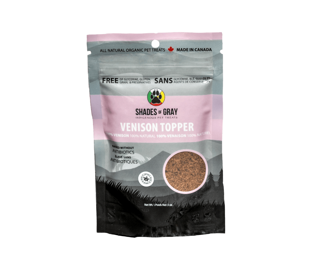 mage of Shades of Gray Indigenous Pet Treats' Venison Topper packaging, a pink bag with a mountain backdrop. Features 100% venison, raised without antibiotics, 100% natural, and free of glycerine, gluten, grain, and preservatives. Includes 'Made in Canada' and 'Ontario' badges, with a Shades of Gray Indigenous Pet Treats paw print circular logo and net weight of 3 oz."